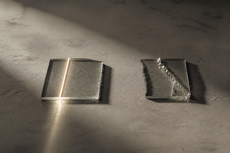 Two small pieces of clear glass on a pale concrete-grey surface — one marked by a single precise line catching the light, the other by an irregular natural-looking fracture — two takes on a deliberate flaw.