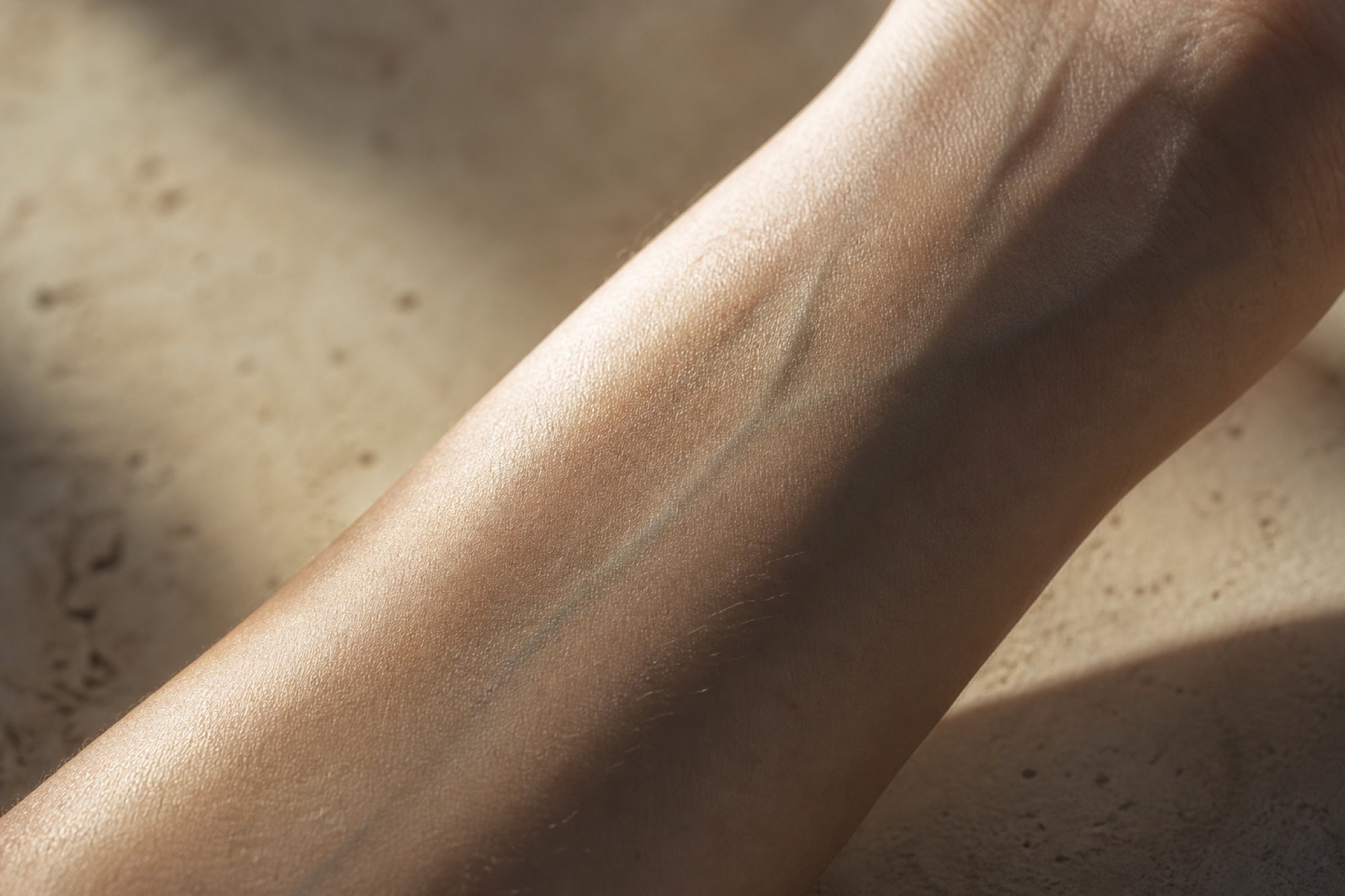 Extreme macro of a bare inner forearm and wrist in warm raking afternoon light on pale concrete, faint vein lines visible — the skin as a living surface that varies from person to person.